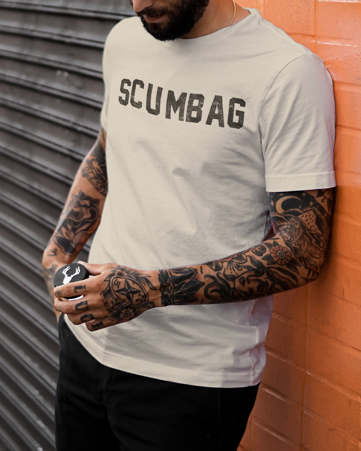 Man in a brown beanie and sunglasses wearing the Scumbag t-shirt in natural with distressed black varsity lettering, leaning against an orange brick wall